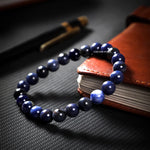 Blue beaded bracelet on a dark textured surface with a book and pen in the background.